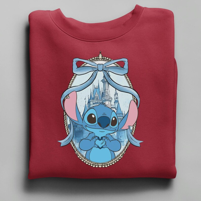 K&ouml;nnte beinhalten: Ein weinrotes Sweatshirt mit einer blauen Cartoonfigur, die ein Herz h&auml;lt, umrahmt von einer hellblauen Schleife und einem Schloss im Hintergrund. Die Figur hat gro&szlig;e Augen und rosa Ohren. Das Design ist auf dem gefalteten Kleidungsst&uuml;ck zentriert.