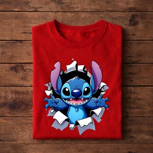 May include: A folded red t-shirt featuring a cartoon illustration of Stitch bursting through a torn hole. The blue character has large ears, a wide grin, and outstretched arms. The background is a wooden surface.