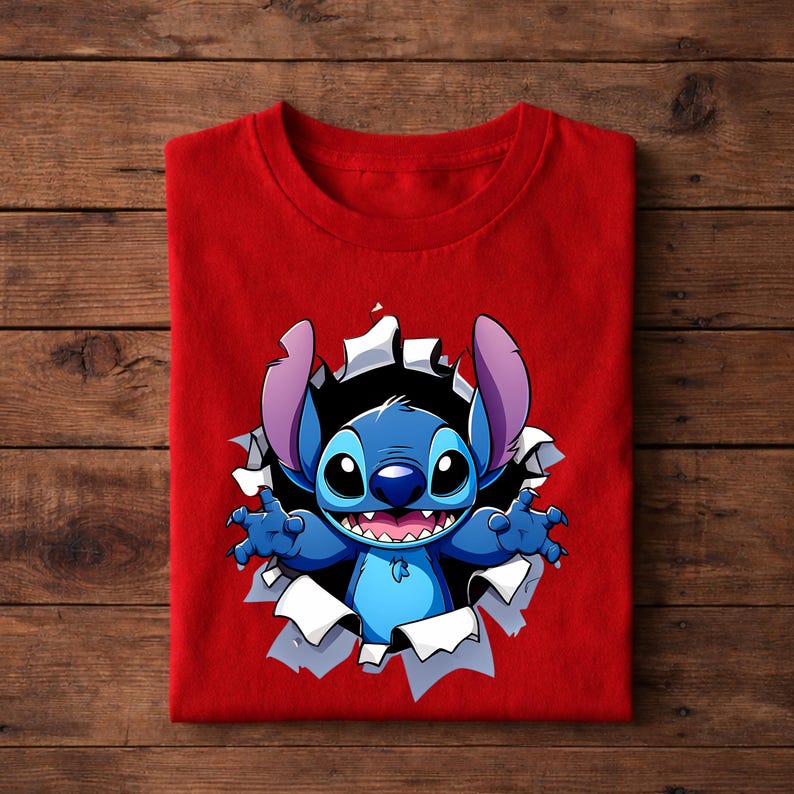 May include: A folded red t-shirt featuring a cartoon illustration of Stitch bursting through a torn hole. The blue character has large ears, a wide grin, and outstretched arms. The background is a wooden surface.