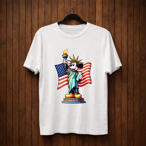 May include: White t-shirt featuring a cartoon character dressed as the Statue of Liberty, holding a torch and a book, with an American flag in the background. The character is standing on a pedestal. The design is colorful and eye-catching.