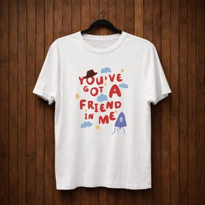 Op de afbeelding: Wit T-shirt met de tekst "You've Got A Friend In Me" in rood, blauw en geel. Het ontwerp bevat een bruine hoed, blauwe wolken, gele sterren en een paarse kwalachtige figuur. Het T-shirt hangt aan een zwarte hanger.