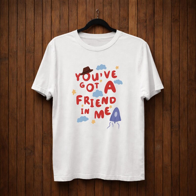 Op de afbeelding: Wit T-shirt met de tekst "You've Got A Friend In Me" in rood, blauw en geel. Het ontwerp bevat een bruine hoed, blauwe wolken, gele sterren en een paarse kwalachtige figuur. Het T-shirt hangt aan een zwarte hanger.