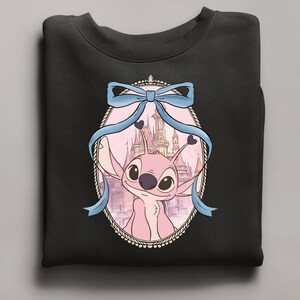 Puede incluir: Sudadera negra con un personaje de dibujos animados rosa en un marco ovalado, sobre un fondo de castillo. Un lazo azul claro adorna la parte superior del marco. La sudadera est&aacute; hecha de un material suave. La imagen es una caricatura.