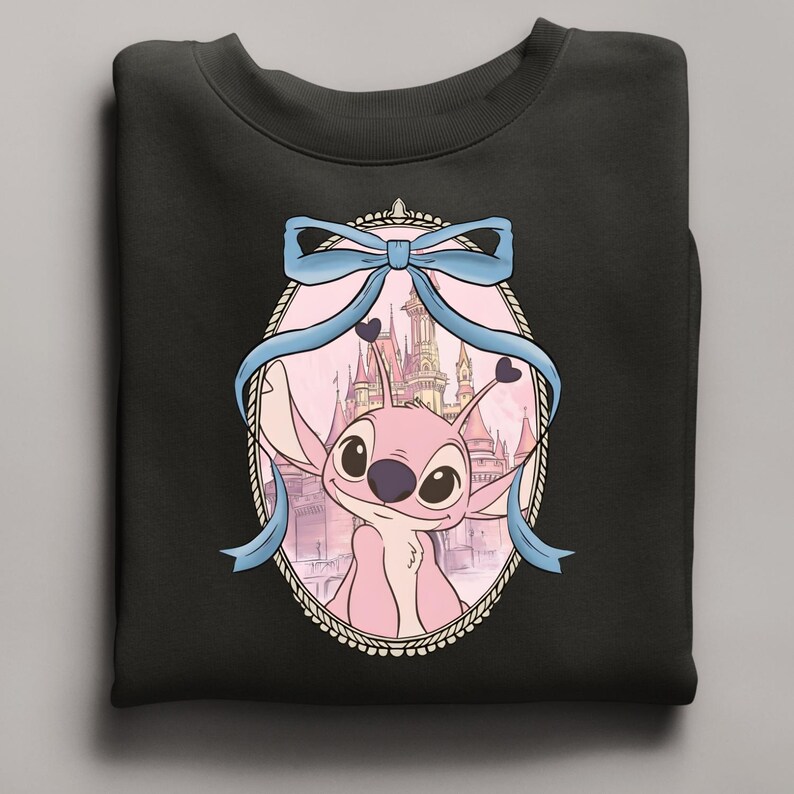 Puede incluir: Sudadera negra con un personaje de dibujos animados rosa en un marco ovalado, sobre un fondo de castillo. Un lazo azul claro adorna la parte superior del marco. La sudadera est&aacute; hecha de un material suave. La imagen es una caricatura.