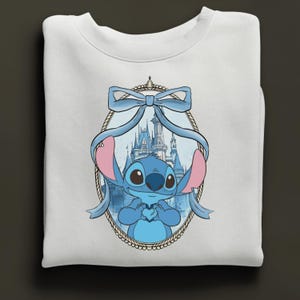 K&ouml;nnte beinhalten: Wei&szlig;es Sweatshirt mit einer blauen Cartoonfigur, die ein Herz h&auml;lt, umrahmt von einer blauen Schleife und einem Schloss im Hintergrund. Die Figur hat gro&szlig;e Augen und rosa Ohren. Das Design ist auf der Vorderseite des gefalteten Sweatshirts zentriert.