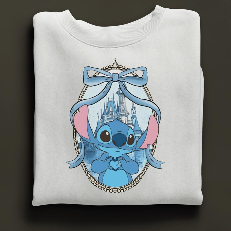 K&ouml;nnte beinhalten: Wei&szlig;es Sweatshirt mit einer blauen Cartoonfigur, die ein Herz h&auml;lt, umrahmt von einer blauen Schleife und einem Schloss im Hintergrund. Die Figur hat gro&szlig;e Augen und rosa Ohren. Das Design ist auf der Vorderseite des gefalteten Sweatshirts zentriert.