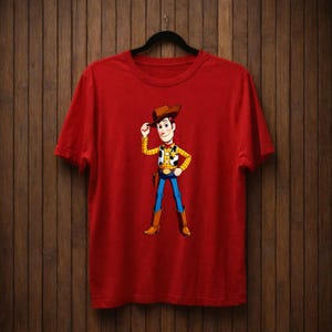 May include: Red t-shirt with a cartoon cowboy character illustration. The cowboy wears a brown hat, yellow shirt, blue trousers, and brown boots. The character is tipping his hat with one hand.