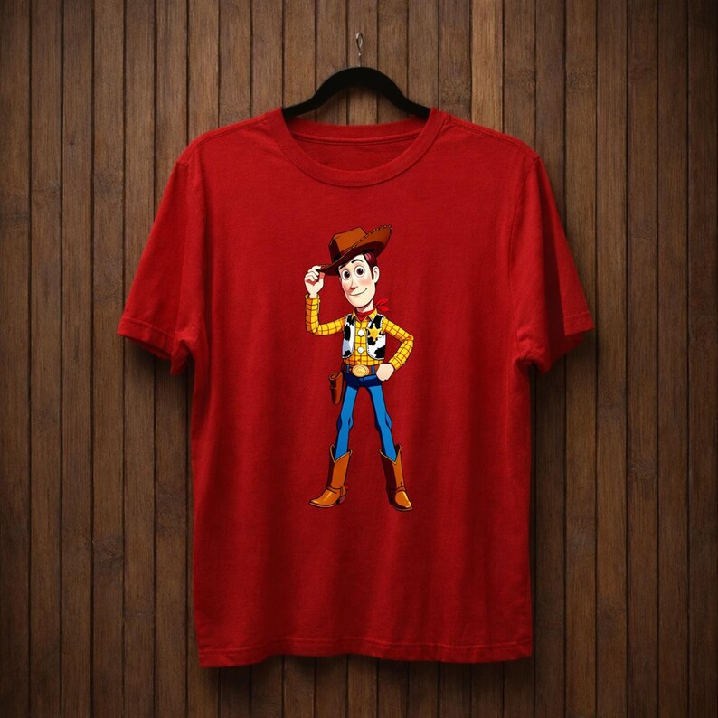 May include: Red t-shirt with a cartoon cowboy character illustration. The cowboy wears a brown hat, yellow shirt, blue trousers, and brown boots. The character is tipping his hat with one hand.