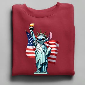 Puede incluir: Una sudadera granate con un personaje de dibujos animados como la Estatua de la Libertad, sosteniendo una antorcha y una tableta, con una bandera estadounidense de fondo. El personaje es azul con ojos grandes y una oreja grande.