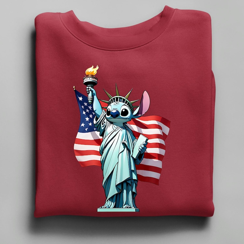 Puede incluir: Una sudadera granate con un personaje de dibujos animados como la Estatua de la Libertad, sosteniendo una antorcha y una tableta, con una bandera estadounidense de fondo. El personaje es azul con ojos grandes y una oreja grande.
