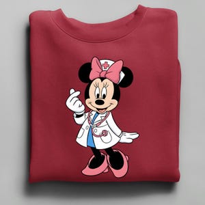 Puede incluir: Una sudadera burdeos con una Minnie Mouse de dibujos animados vestida de enfermera. Minnie lleva una bata blanca, un lazo rosa y un estetoscopio. Sus manos forman un coraz&oacute;n. El dise&ntilde;o est&aacute; centrado en la sudadera doblada.