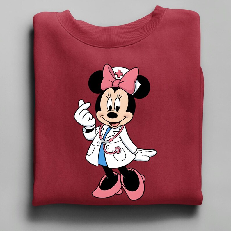 Puede incluir: Una sudadera burdeos con una Minnie Mouse de dibujos animados vestida de enfermera. Minnie lleva una bata blanca, un lazo rosa y un estetoscopio. Sus manos forman un coraz&oacute;n. El dise&ntilde;o est&aacute; centrado en la sudadera doblada.