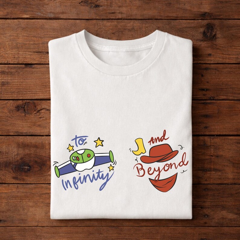 Op de afbeelding: Wit T-shirt met de kleurrijke tekst "To Infinity and Beyond". Het ontwerp bevat een cartoon ruimteschip en een cowboyhoed met een rode bandana. Het shirt is op een houten oppervlak gevouwen.