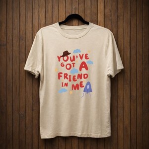 Op de afbeelding: Cr&egrave;mekleurig T-shirt met de tekst "You've Got A Friend In Me" in rood en blauw, met een bruine hoed, blauwe wolken en gele sterren. Het shirt hangt aan een zwarte hanger tegen een houten achtergrond.