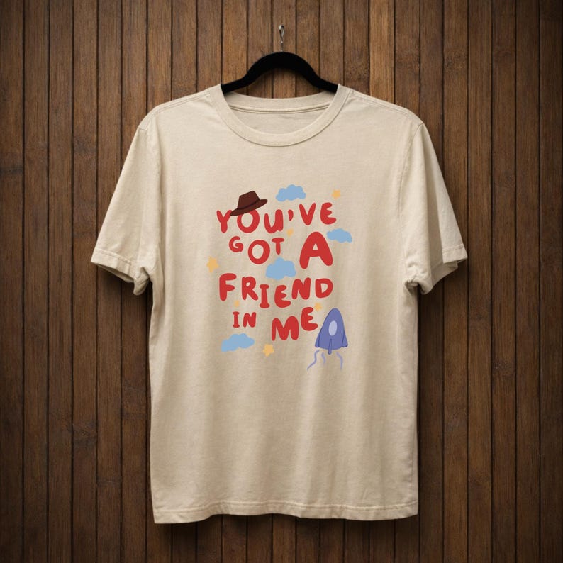 Op de afbeelding: Cr&egrave;mekleurig T-shirt met de tekst "You've Got A Friend In Me" in rood en blauw, met een bruine hoed, blauwe wolken en gele sterren. Het shirt hangt aan een zwarte hanger tegen een houten achtergrond.