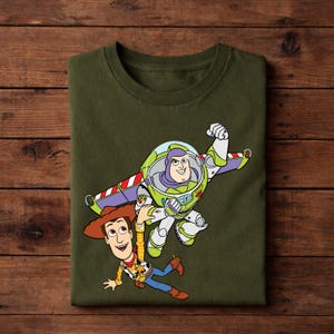 May include: An olive green t-shirt featuring a colorful graphic of Buzz Lightyear and Woody from Toy Story. Buzz is in his space suit, and Woody is wearing his cowboy hat and vest. The design is a cartoon style.