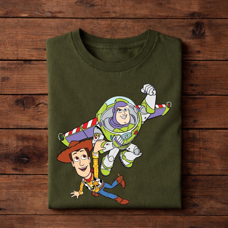 May include: An olive green t-shirt featuring a colorful graphic of Buzz Lightyear and Woody from Toy Story. Buzz is in his space suit, and Woody is wearing his cowboy hat and vest. The design is a cartoon style.