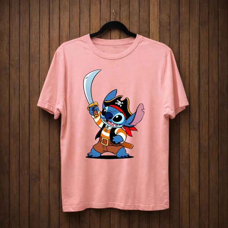 May include: A light pink t-shirt featuring a cartoon character dressed as a pirate, holding a sword. The character is blue with large eyes, wearing a pirate hat, vest, and red bandana. The shirt is hanging on a black hanger.