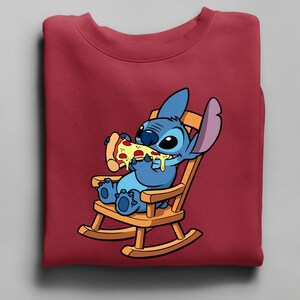 Pu&ograve; includere: Una felpa bordeaux con un'illustrazione di Stitch, un alieno blu, che mangia una fetta di pizza su una sedia a dondolo di legno. La pizza ha formaggio giallo e peperoni rossi. La felpa &egrave; piegata.
