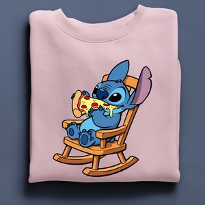 Pu&ograve; includere: Una felpa rosa chiaro con un'illustrazione di Stitch di Lilo & Stitch. Stitch, in blu, &egrave; seduto su una sedia a dondolo di legno e mangia una pizza. La pizza ha formaggio giallo e peperoni rossi.