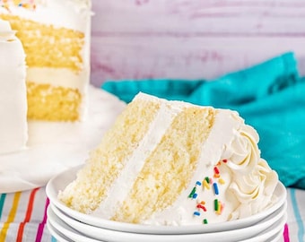 May include: A slice of layered yellow cake with white frosting and colorful sprinkles sits on a white plate. The cake has a creamy frosting between the layers and on top, with a decorative swirl.
