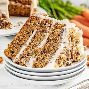 Puede incluir: Una porción de pastel de zanahoria con glaseado blanco y nueces picadas, servida en una pila de platos blancos. El pastel tiene varias capas con glaseado entre cada capa. Zanahorias y vegetación son visibles en el fondo.