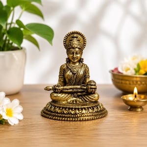May include: A metallic gold-toned statue of a seated deity figure, featuring intricate carvings on the clothing, jewelry, and crown. The figure sits in a cross-legged position on a circular, ornate pedestal base with a lotus petal design.