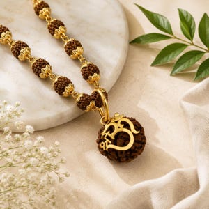 以下が含まれることがあります： 茶色で質感のあるルドラクシャビーズを金色の金属キャップで区切ったネックレス。ペンダントは、金色のオームのシンボルが施された大きめのルドラクシャビーズで構成され、金色の金属製の丸いリングから吊り下げられています。