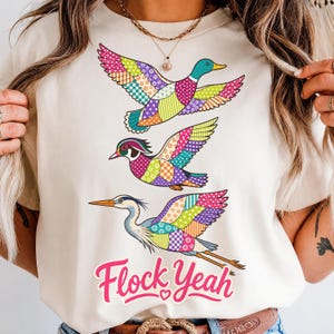 Puede incluir: Una camiseta de color crema con un gráfico de tres aves volando, cada una decorada con patrones de patchwork coloridos en tonos rosa, verde, naranja y morado. Debajo de las aves, el texto Flock Yeah está impreso en una fuente rosa llamativa.
