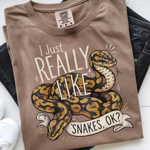 May include: A brown t-shirt featuring a detailed illustration of a snake with yellow and black markings. The text "I Just REALLY LIKE SNAKES, OK?" is printed above and below the snake in a playful font. The shirt is folded.