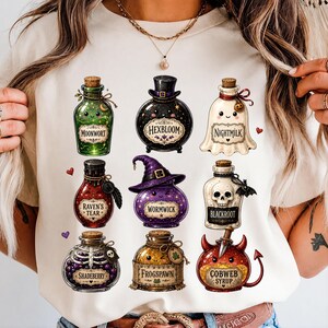 May include: A cream-colored t-shirt featuring a graphic print of nine distinct potion bottles with faces. The bottles are labeled Moonwort, Hexbloom, Nightmilk, Raven's Tear, Wormwick, Blackroot, Shadeberry, Frogspawn, and Cobweb Syrup.