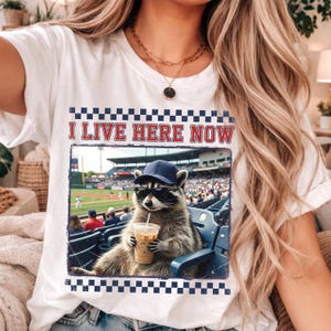 Può includere: T-shirt bianco con la scritta "I LIVE HERE NOW" in lettere rosse. La maglietta presenta una grafica di un procione con occhiali da sole e un berretto da baseball, che tiene in mano una bevanda al caffè e seduto in uno stadio.