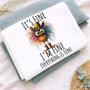 May include: White folded fabric with a whimsical illustration of a colorful creature wearing a cowboy hat. The text "IT'S FINE, I'M FINE, EVERYTHING IS FINE" is printed in black. The fabric is folded on top of a light blue fabric.