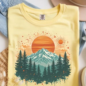 May include: Yellow t-shirt featuring a mountain landscape design. The graphic includes a large orange sun, a smaller sun, a mountain range, and evergreen trees. The shirt has a crew neck and short sleeves.