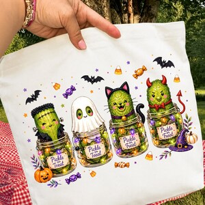 Könnte beinhalten: Eine weiße Tragetasche mit einem Grafikdruck von vier grünen Gewürzgurken in Gläsern. Die Gurken sind als Frankenstein-Monster, Geist, Katze und Teufel verkleidet. Jedes Glas hat ein Etikett mit der Aufschrift Pickle or Treat, ergänzt durch Fledermäuse, Süßigkeiten und Kürbisse.