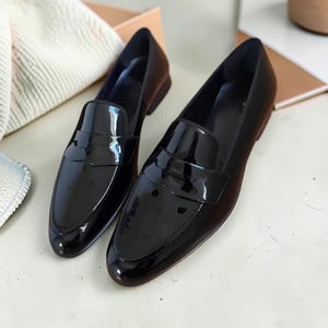 May include: A pair of black patent leather loafers featuring a glossy finish, a rounded toe, and a classic strap across the vamp. The shoes have a low heel and a smooth, reflective surface that catches the light, showcasing a clean silhouette.