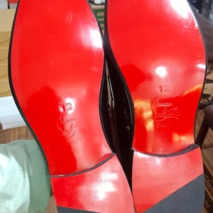May include: A pair of black dress shoes with glossy red soles. The soles feature the embossed text 'Christian Louboutin Paris' and the number '12' in the center. Each heel has a black textured rubber tap at the bottom for traction.