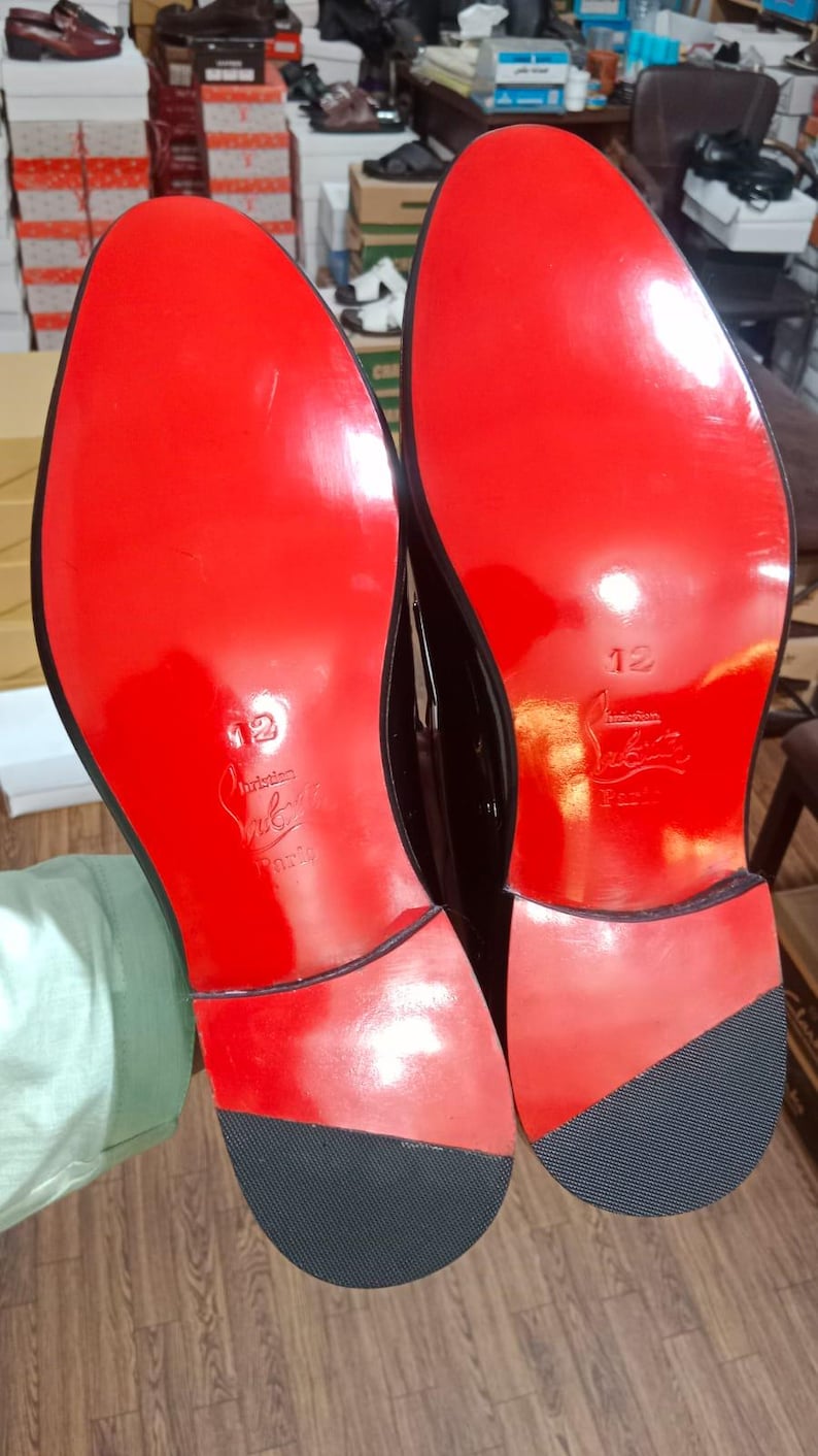 May include: A pair of black dress shoes with glossy red soles. The soles feature the embossed text 'Christian Louboutin Paris' and the number '12' in the center. Each heel has a black textured rubber tap at the bottom for traction.