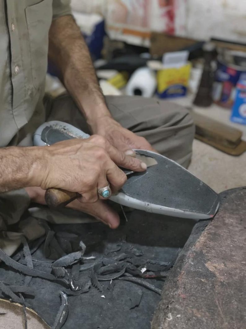 May include: A person uses a sharp metal tool with a wooden handle to trim the edges of a gray shoe sole. The sole is held firmly while excess dark material is shaved away, leaving small scraps of material scattered on the work surface below.