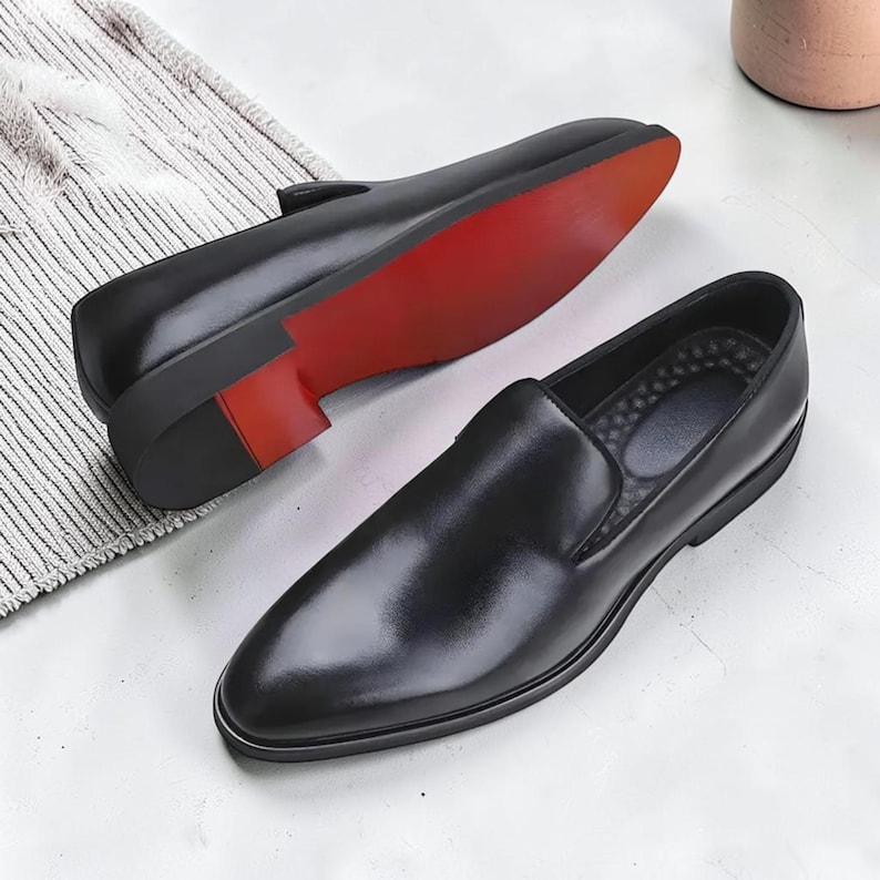 K&ouml;nnte beinhalten: Ein Paar schwarze Slipper mit glatter Oberfl&auml;che und spitzer Zehenpartie. Ein Schuh ist so positioniert, dass das Innere sichtbar ist, w&auml;hrend der andere umgedreht ist, um eine leuchtend rote Sohle und einen niedrigen Blockabsatz zu zeigen.
