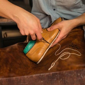 Pode incluir: Um sapato oxford de couro castanho-claro com detalhes decorativos brogue sendo costurado com uma agulha e fio branco. O sapato apresenta uma sola de cor clara e uma inser&ccedil;&atilde;o azul-petr&oacute;leo vis&iacute;vel na abertura do calcanhar sobre couro castanho.