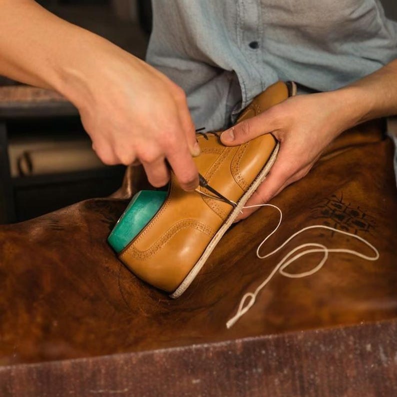 Pode incluir: Um sapato oxford de couro castanho-claro com detalhes decorativos brogue sendo costurado com uma agulha e fio branco. O sapato apresenta uma sola de cor clara e uma inser&ccedil;&atilde;o azul-petr&oacute;leo vis&iacute;vel na abertura do calcanhar sobre couro castanho.