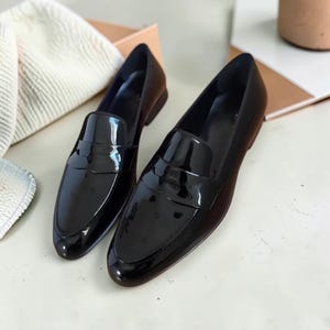 May include: A pair of black patent leather loafers featuring a glossy finish, a rounded toe, and a classic strap across the vamp. The shoes have a low heel and a smooth, reflective surface that catches the light, showcasing a clean silhouette.