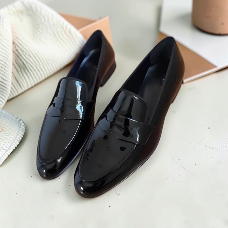 May include: A pair of black patent leather loafers featuring a glossy finish, a rounded toe, and a classic strap across the vamp. The shoes have a low heel and a smooth, reflective surface that catches the light, showcasing a clean silhouette.