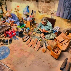 K&ouml;nnte beinhalten: Zwei Personen arbeiten in einer Werkstatt, umgeben von Lederschuhsohlen, Werkzeugen und Materialien. Eine Person sitzt auf dem Boden und fertigt einen Schuh an, w&auml;hrend eine andere in der N&auml;he arbeitet. Der Raum ist mit verschiedenen Holzteilen und Resten gef&uuml;llt.