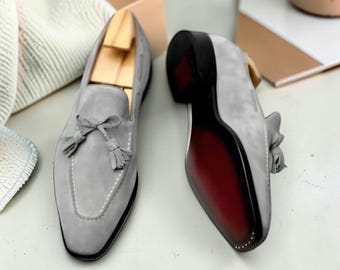 Pode incluir: Um par de mocassins de camurça cinza-claro com uma borla decorativa no peito do pé, costuras brancas em contraste ao longo da biqueira e uma sola escura com o centro vermelho profundo. Um sapato é visto de cima e o outro de baixo.