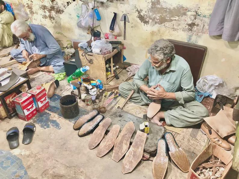 May include: Two shoemakers work in a workshop, crafting footwear. One person sits on the floor shaping a leather shoe sole, while the other works nearby. Various leather pieces, shoe soles, tools, and red cans labeled CEMENTEX are visible.
