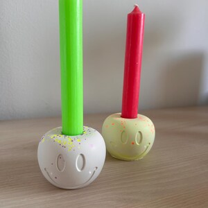 May include: Two decorative candle holders with smiley faces, each holding a tall, thin candle. One candle is bright green, the other is red. The holders are white and yellow with colorful speckles.