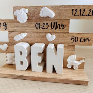 Könnte beinhalten: Ein Baby-Ankündigungsschild aus Holz mit dem Namen "BEN" in weißen Blockbuchstaben. Das Schild enthält dekorative Elemente wie einen Babyfußabdruck, einen Strampler, eine Flasche und einen Kinderwagen. Es zeigt auch das Geburtsdatum, die Uhrzeit, das Gewicht (3.210 g) und die Größe (50 cm) des Babys.