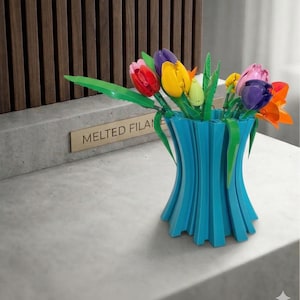 Op de afbeelding: Een kleurrijk boeket tulpen in een turquoise vaas. De tulpen zijn rood, geel, paars, roze en oranje, met groene bladeren. De vaas heeft een uniek, modern design. De tekst "MELTED FILAME" is zichtbaar op een klein bordje.